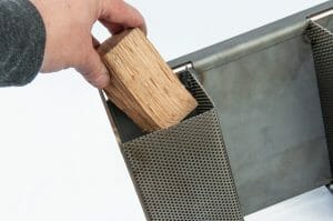 Hand placing wood into the perforated cavity of the Heavy d Stick burning heat diffuser, adding fuel for a smoky and flavorful cooking experience